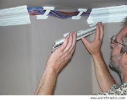 Man installing a white cable raceway with exposed colored wires along a wall ceiling line
