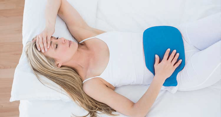 Mulher com dor abdominal deitada na cama com uma bolsa de água quente azul sobre o baixo ventre.