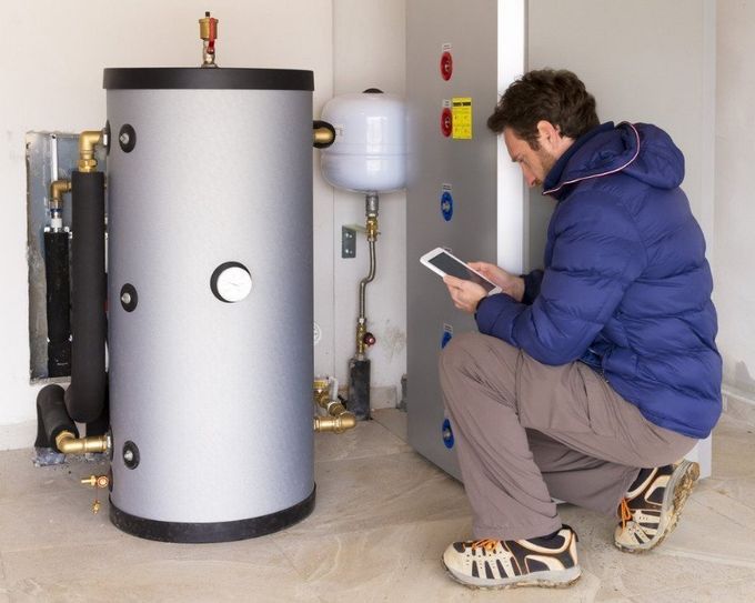 A man is kneeling down in front of a heater and looking at a tablet.