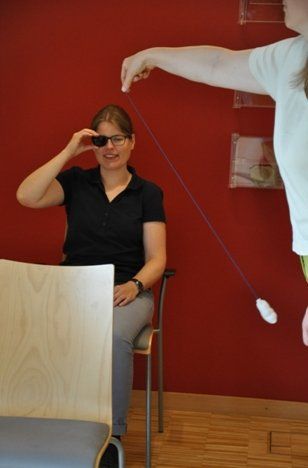 eine Person hält sich ein getöntes Brillenglas einer Sonnenbrille vor ein Auge, eine andere Person schwingt einen Gegenstand an einer Schnur um einen Stuhl vor der ersten Person