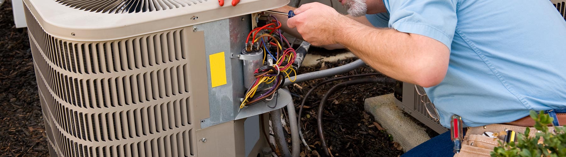 A person in a blue shirt repairs an air conditioning unit outdoors.