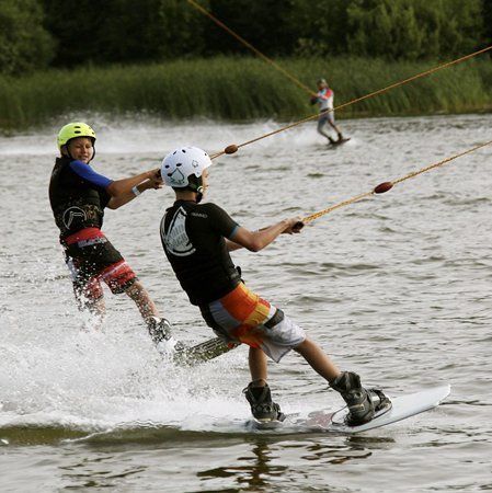 Two wakeboarders being pulled by a cable across a lake. One wears a blue shirt, the other a black shirt.