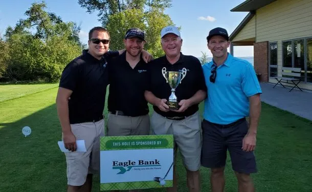 Four people stand outdoors on a golf course holding a trophy behind a tournament sign for Eagle Bank.