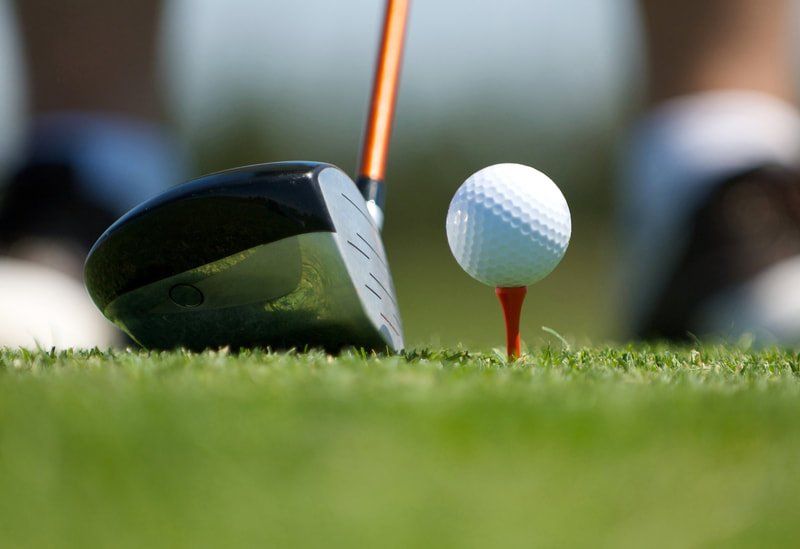 A golf club head is positioned on green grass behind a white golf ball resting on a red tee.