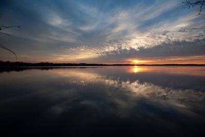 Sunset over a calm, reflective lake with a soft, streaky sky and distant dark treeline.