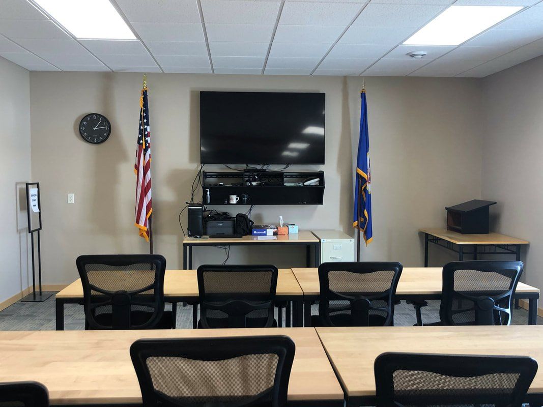 A conference room with two rows of chairs facing a wall-mounted TV, two flags, a clock, and tables.