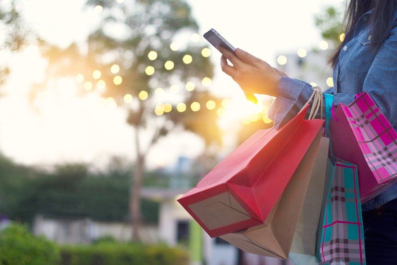 A person in a denim jacket holding a smartphone and several colorful shopping bags against a blurred, sunny park backdrop.