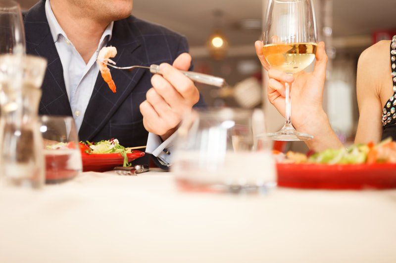 A person in a blazer holds a fork with a shrimp, while another person holds a glass of white wine at a dinner table.