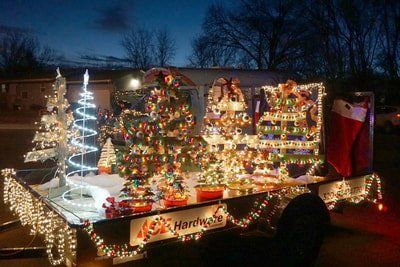 A brightly lit parade float decorated with several illuminated Christmas trees and a large stocking at twilight.