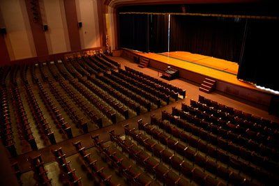 A high-angle view of an empty theater with rows of dark seats facing an illuminated wooden stage with black curtains.