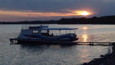 A white and blue pontoon boat is docked on a calm lake at sunset, with the orange sun reflecting on the water.