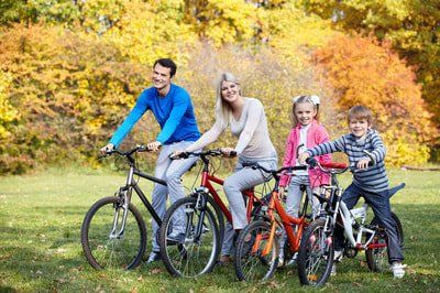 A smiling family of four riding bicycles through a park with autumn foliage in the background.