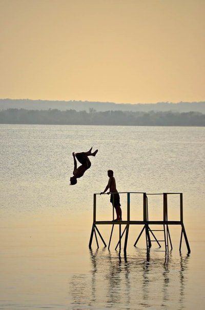 A person performs a backflip off a wooden diving platform into a calm lake during a golden sunset.
