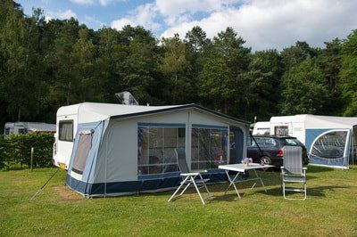 A travel trailer with an attached awning, chairs, and a table set up on a grassy campsite against a backdrop of trees.