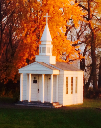 A white wooden chapel with a steeple sits before a backdrop of autumn trees with vibrant orange and gold leaves.