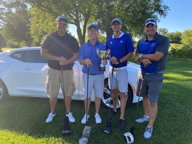 Four people stand in front of a white sports car on a golf course, holding golf clubs and a trophy.