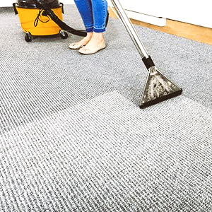 Carpet cleaning with a vacuum nozzle on gray carpet beside a yellow machine and a person’s bare feet