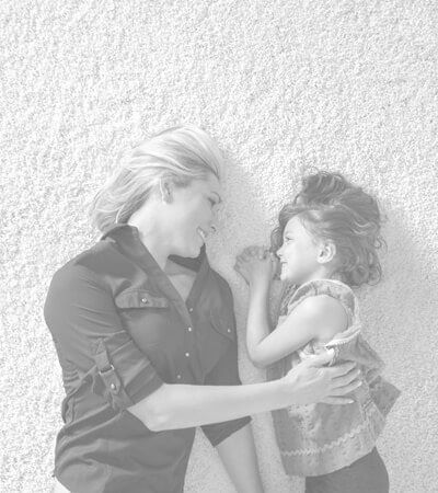 Woman and young girl lying together, smiling and touching noses on a light textured surface.