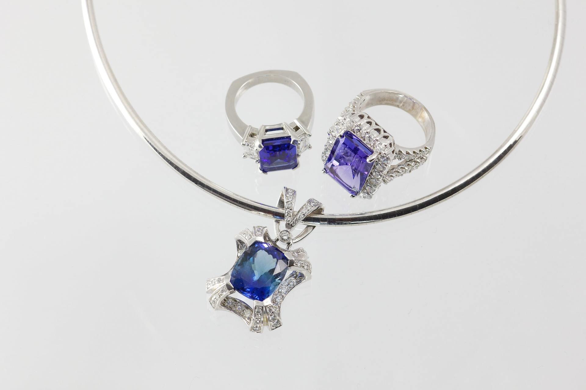 Jewelry set: a sapphire pendant on a silver necklace, with two sapphire rings, set on a reflective surface.