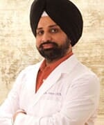 Man with black turban and beard in a white lab coat, arms crossed, smiling.