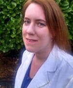 Woman with auburn hair smiles, wearing a blue shirt and white blazer, in front of green bushes.
