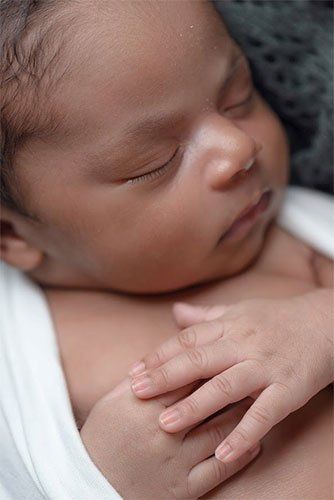 Newborn baby sleeping, dark skin, wrapped in white, hand resting on chest.