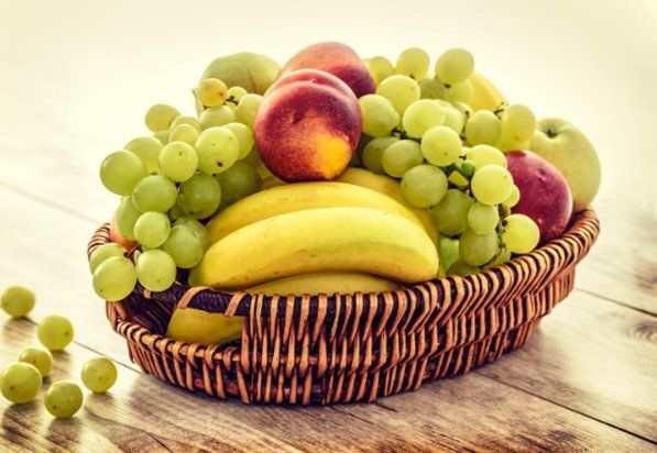 Basket overflowing with green grapes, bananas, peaches, and apples on a wooden surface.