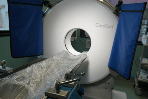A white CoreTom CT scanner with a patient table, blue shields, and a circular opening.