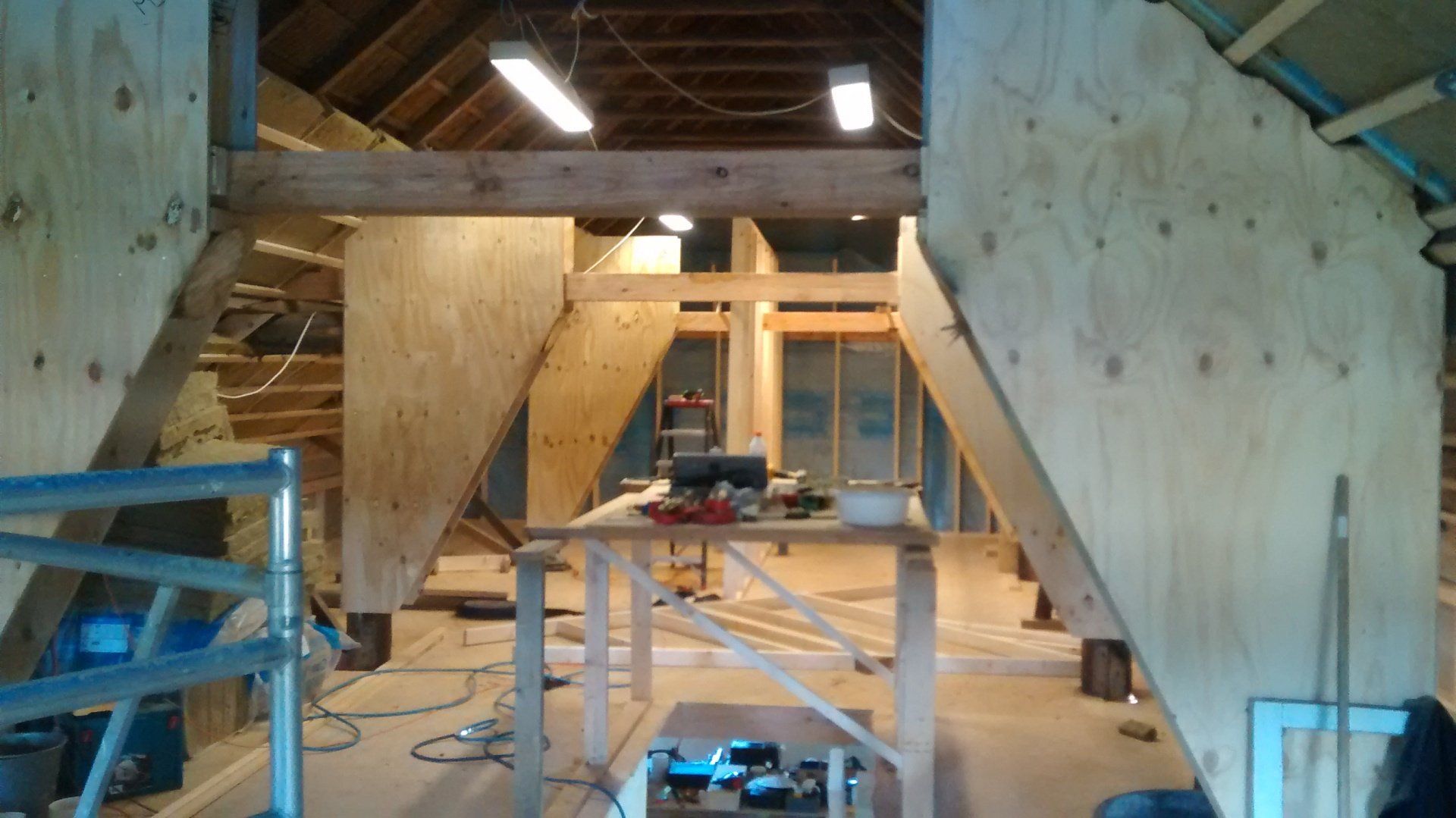 Houtwerkplaats met grote multiplexpanelen in aanbouw, gereedschap op een werkbank en blauwe ladders aan de linkerkant.