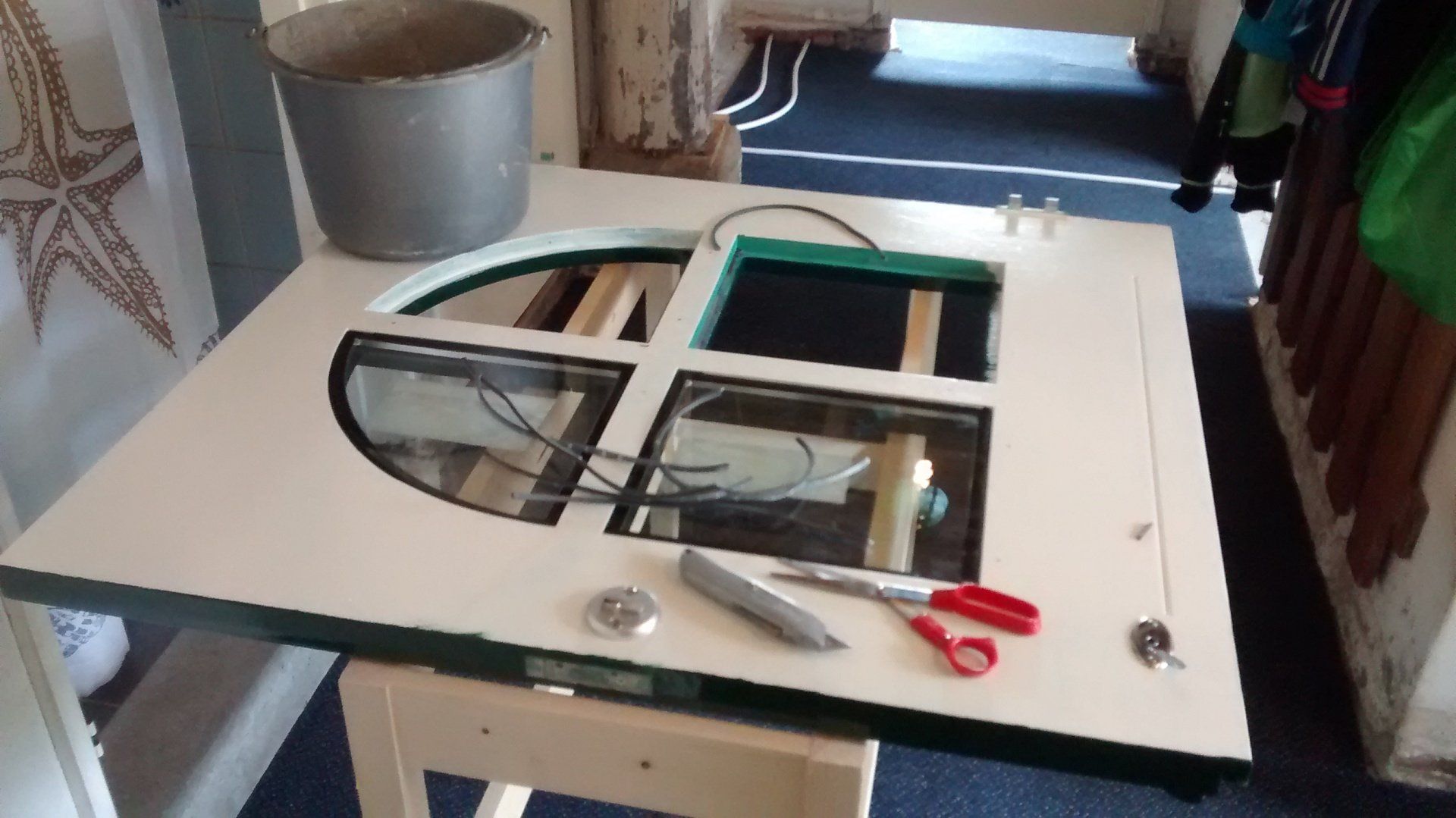 Witte tafel met uitgesneden vormen, gereedschap en een rode schaar in een werkplaatsomgeving.