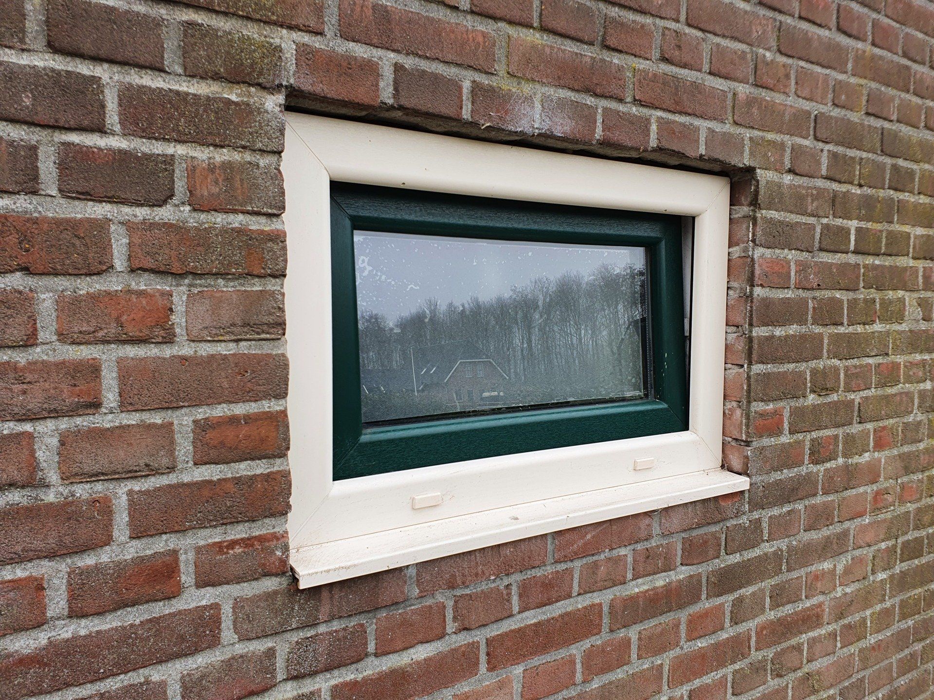 Klein raam met groene kozijnen en witte randen, ingebouwd in een bakstenen muur.