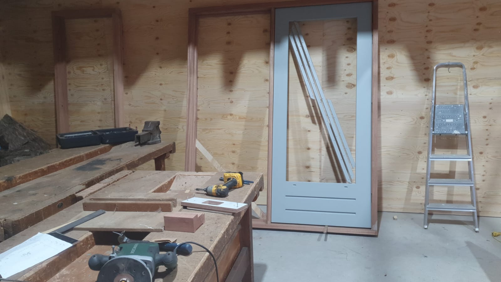 Werkplaatsinterieur met houten frame, werkbank, deur, ladder en gereedschap op de vloer.