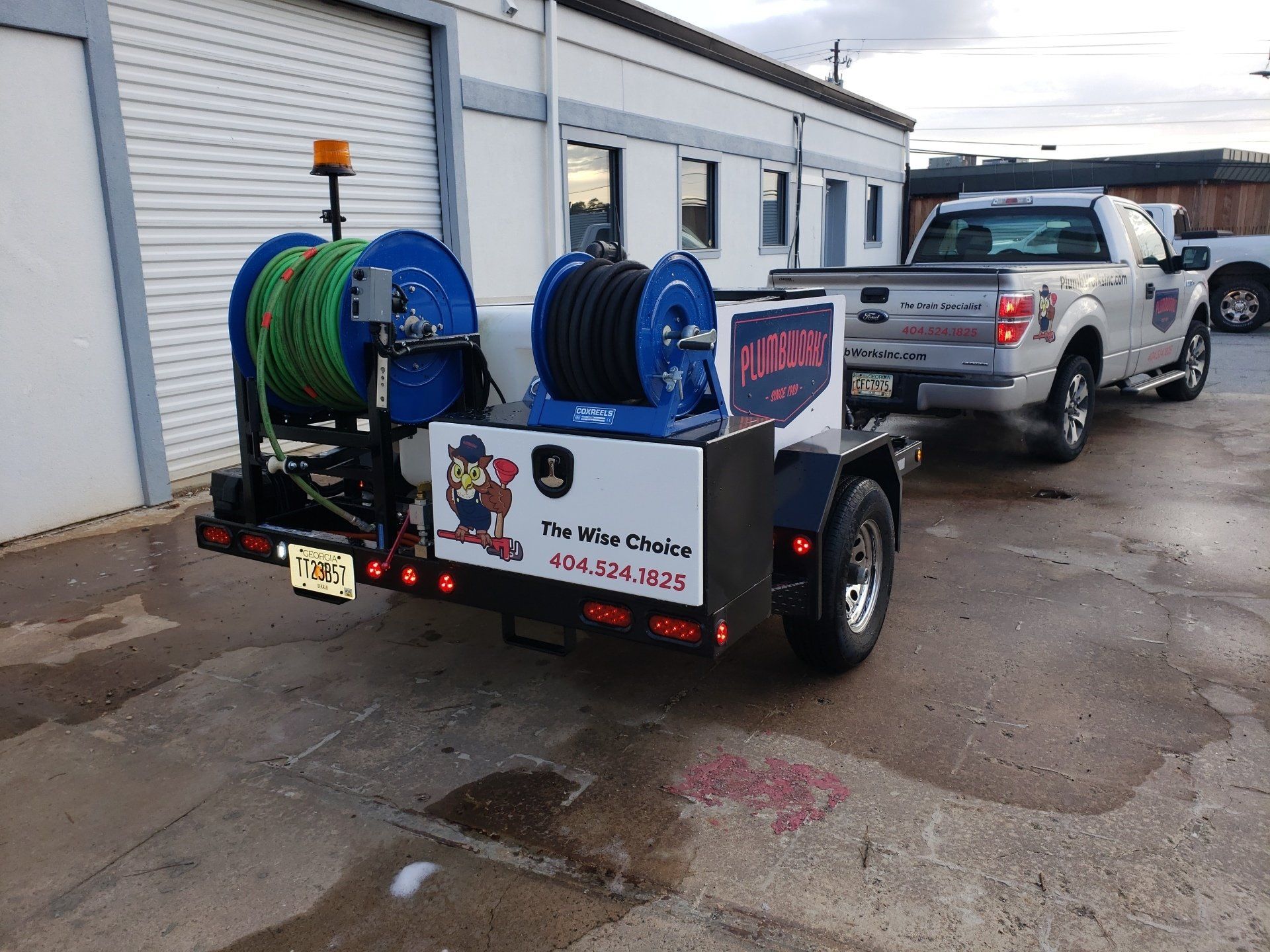 Truck pulling a trailer with two large hoses, a light, and company logo in front of a building.