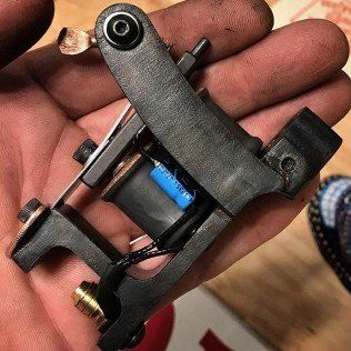 Black tattoo machine with blue coil held in a hand, close-up on a wooden surface