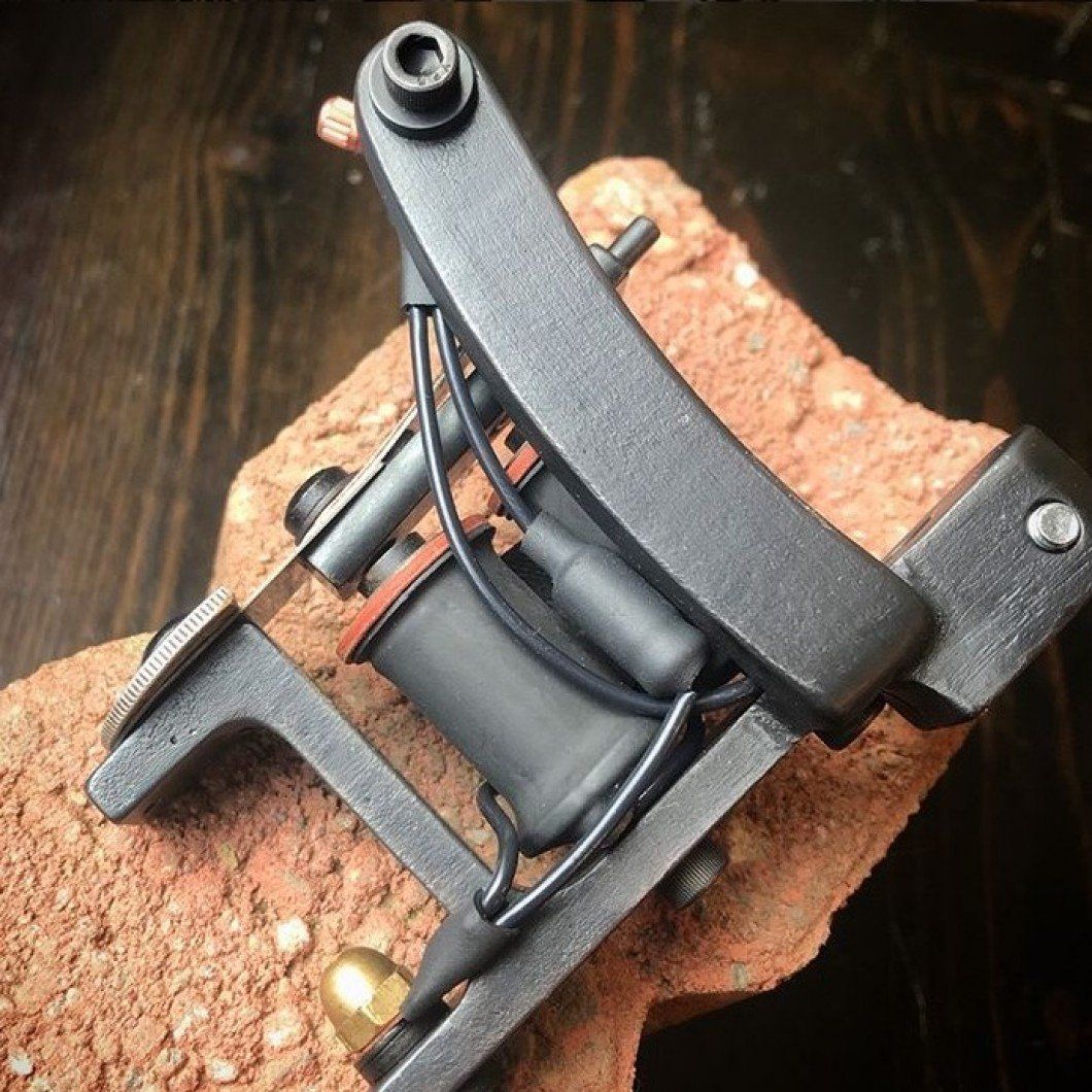 Small black tattoo machine with red and black wires resting on a cork block.