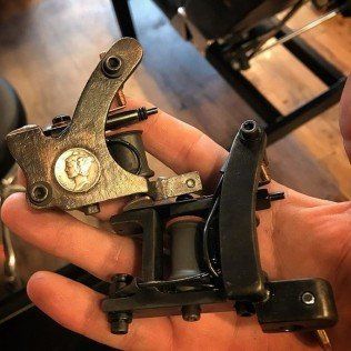 Hand holding two black and brass tattoo machine frames on a wooden floor.