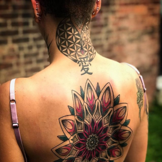 Back of a person with a large colorful floral tattoo and geometric neck tattoo, against a brick wall