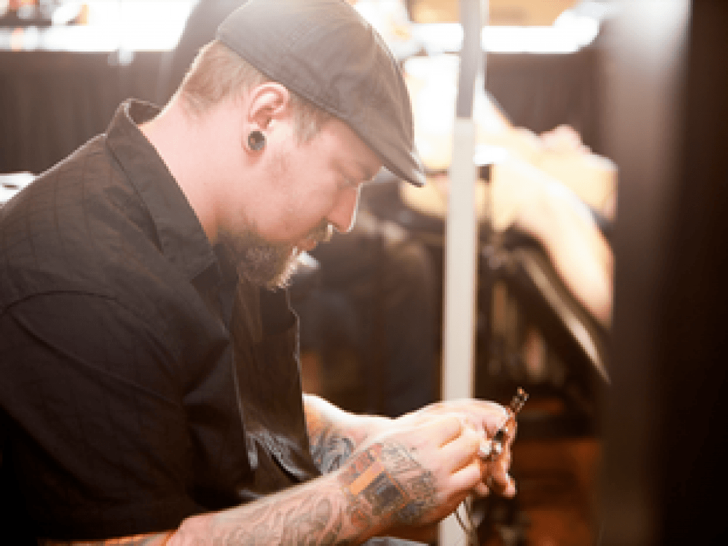 Person in a black shirt and cap leaning forward while working on a tattoo in a warmly lit studio