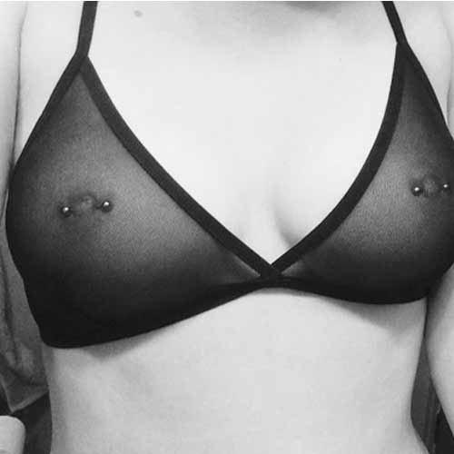 Person wearing a black sheer bralette with silver nipple piercings against a plain background