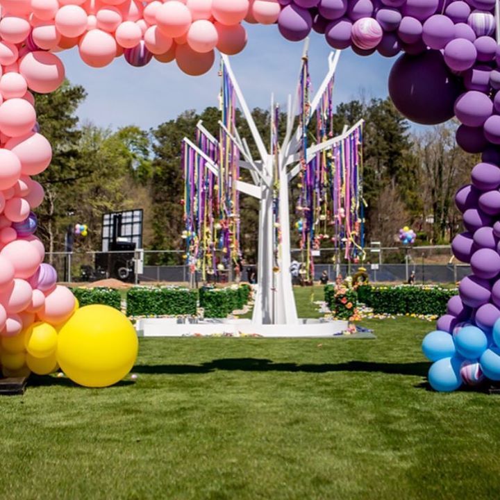A multi color balloon garland welcomes guests as they approach a man made tree.