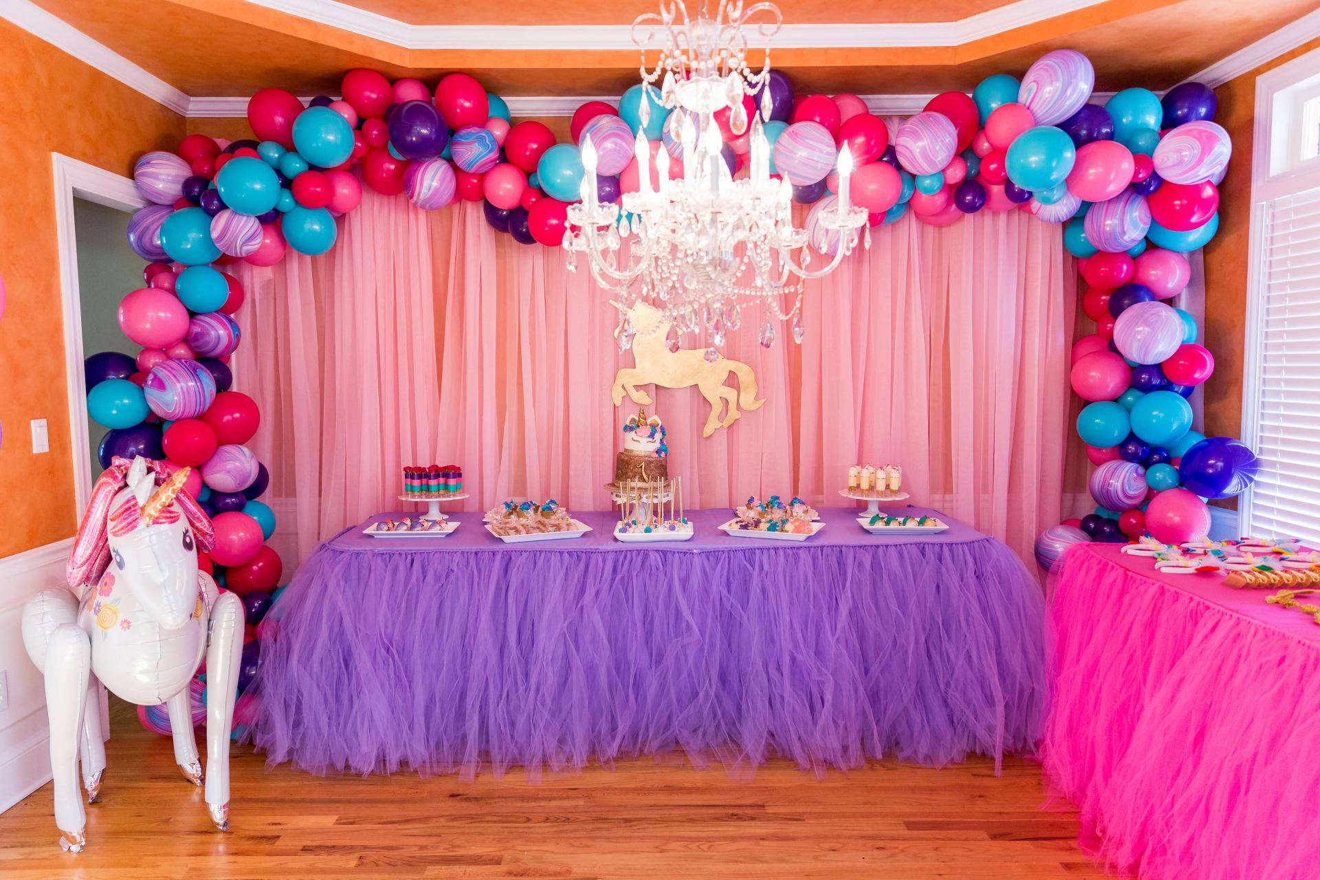 Colorful unicorn themed first birthday dessert table with balloon arch and chandelier backdrop