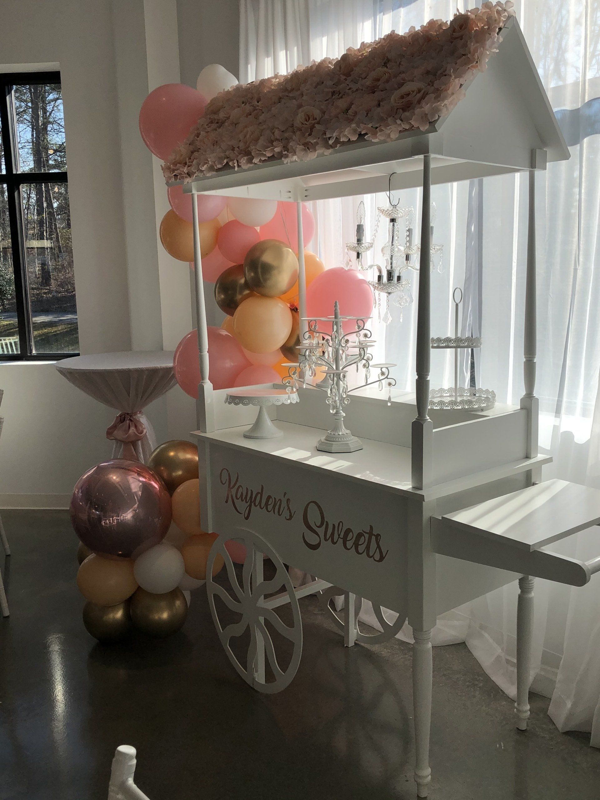 White dessert cart rental with floral canopy and peach gold balloon décor for parties