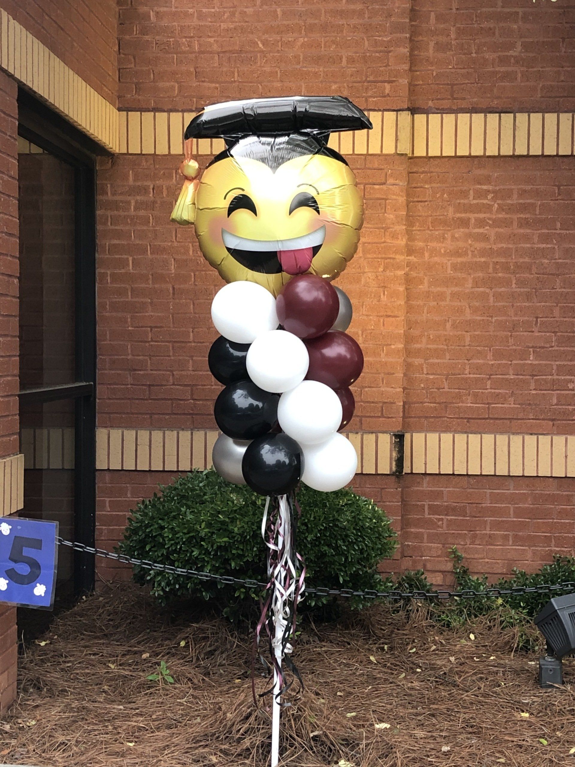 Graduation emoji balloon with cap and tassel styled on balloon column in front of brick wall