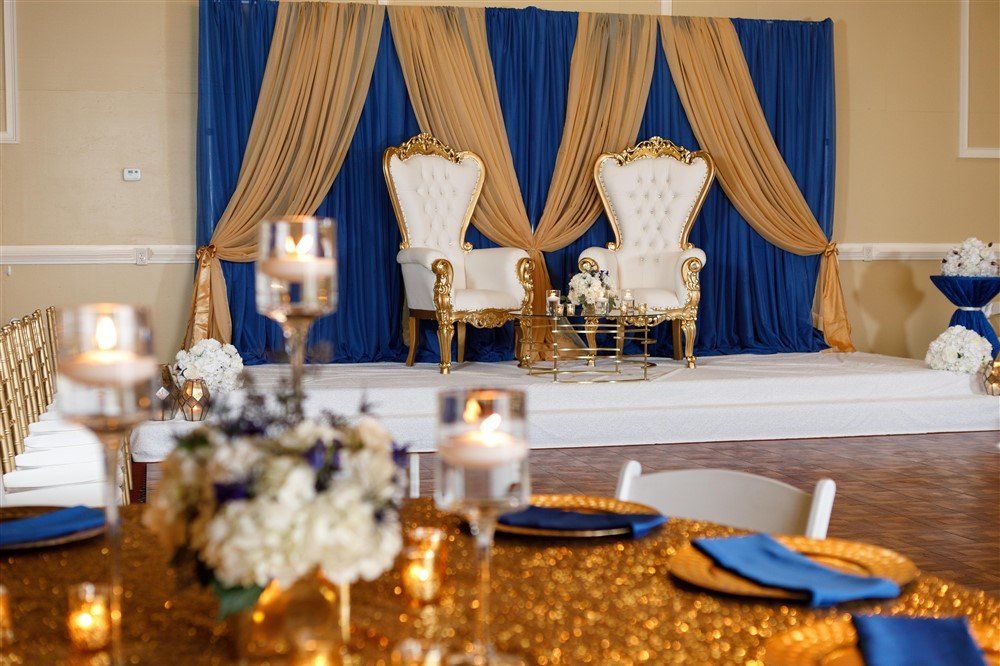 Royal blue and gold stage backdrop with twin throne chairs and draped fabric for wedding or milestone event head table