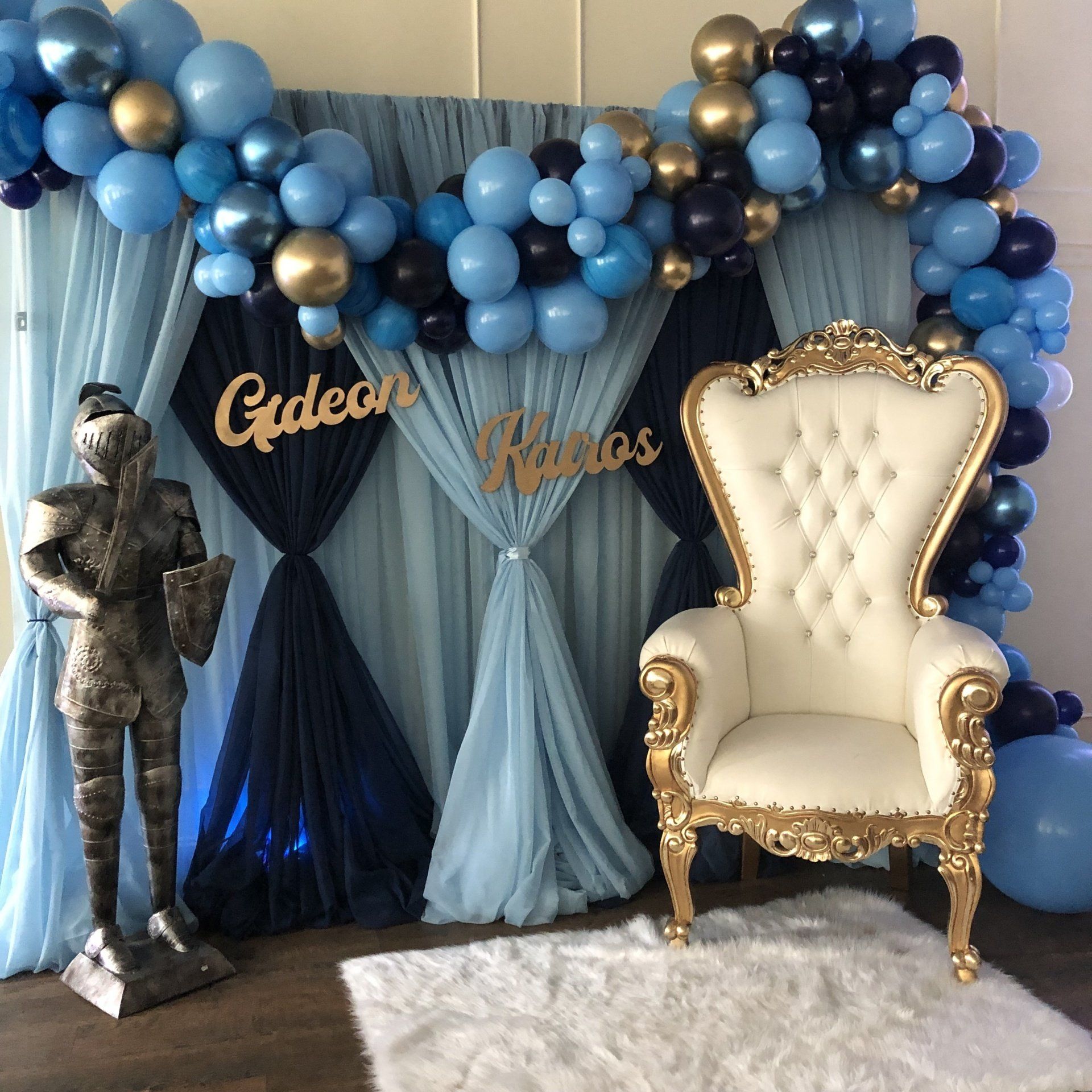 Blue and gold balloon arch with custom name sign and white throne chair for baby shower photo backdrop