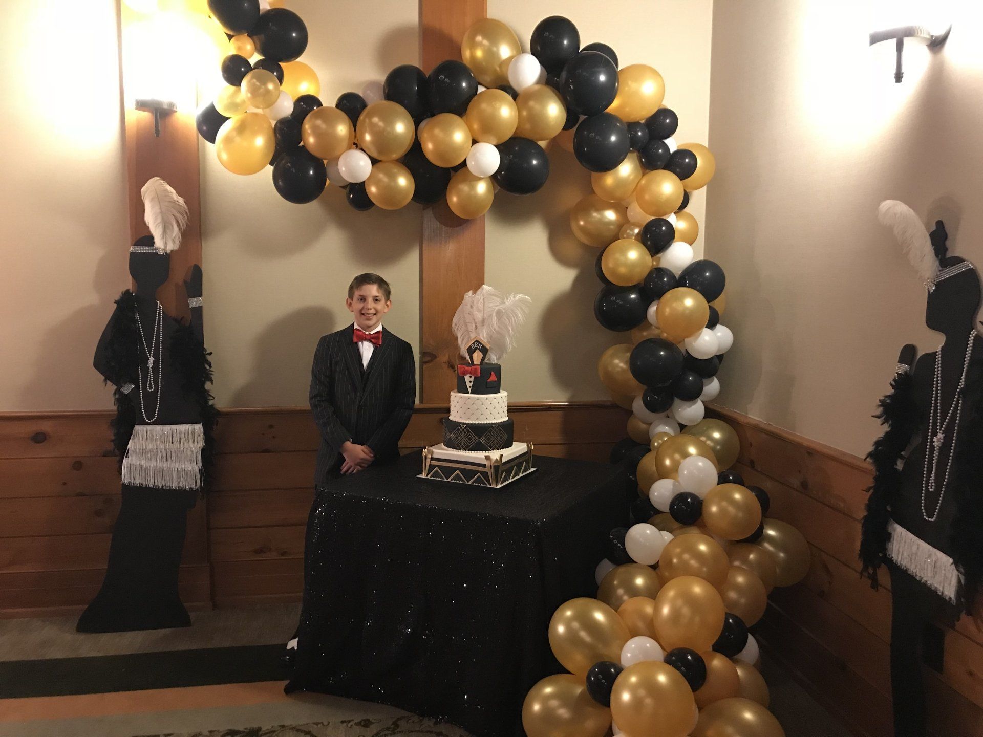 A young gentleman stands next to a cake table decorated in the roaring 20's style and a beautiful balloon garland.