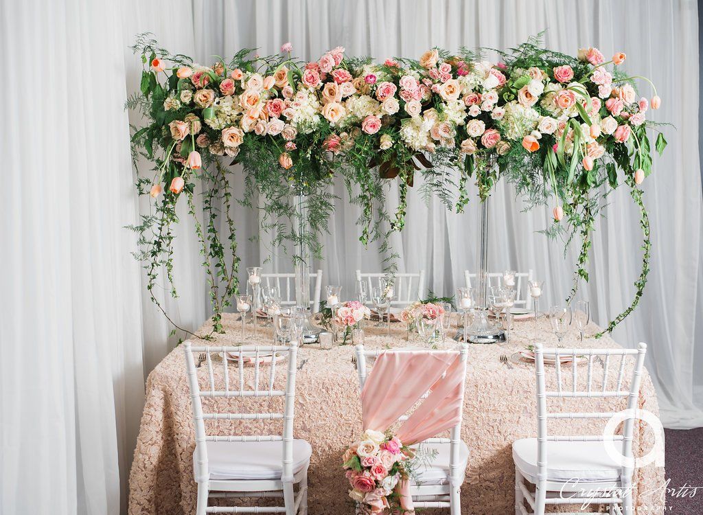 a table setup with an elevated bridge floral centerpiece, candles, and white banquet chairs.