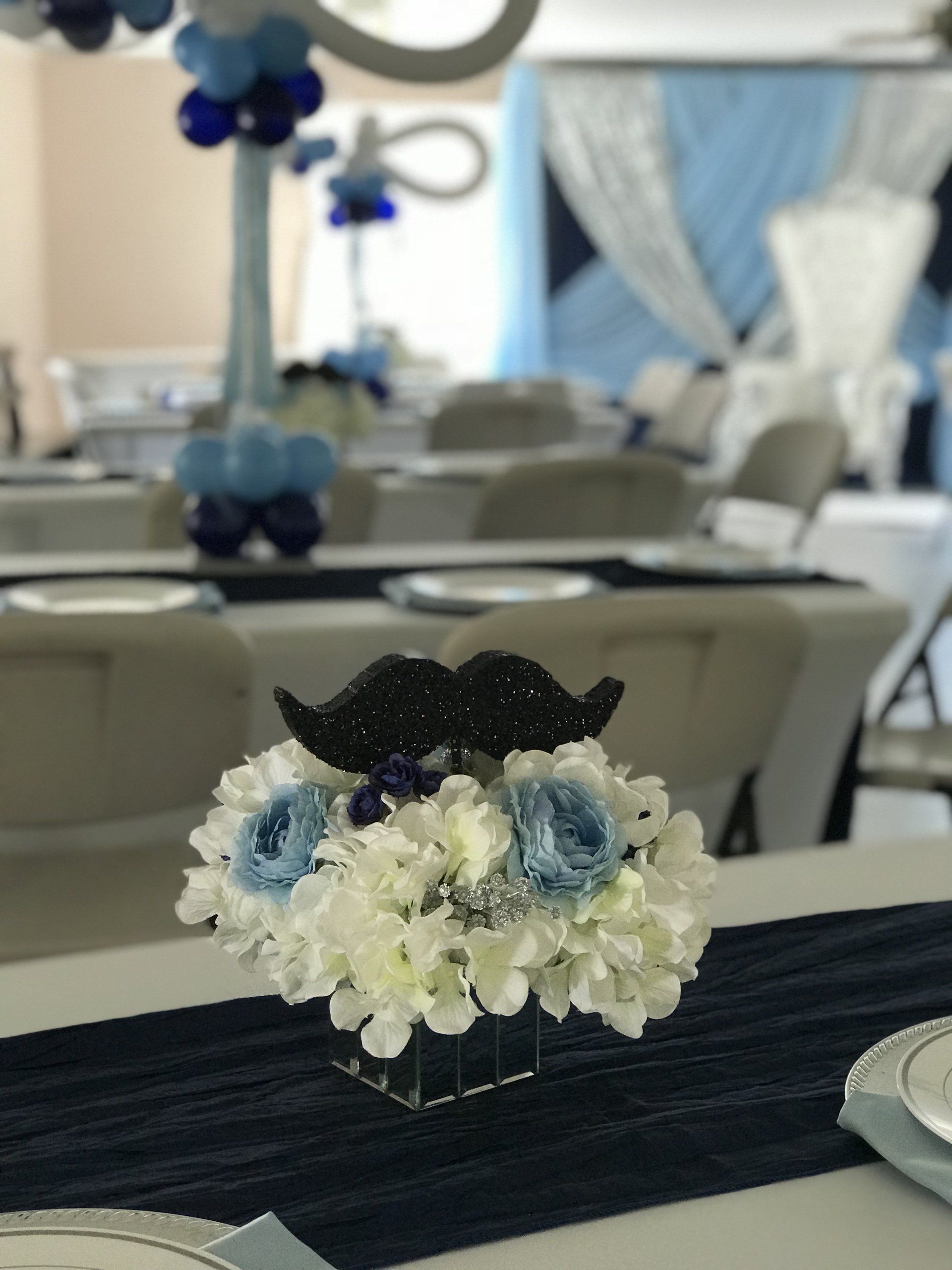 Small floral centerpiece in a mirror cube base and a sparkling black mustache prop at the top for a baby shower.