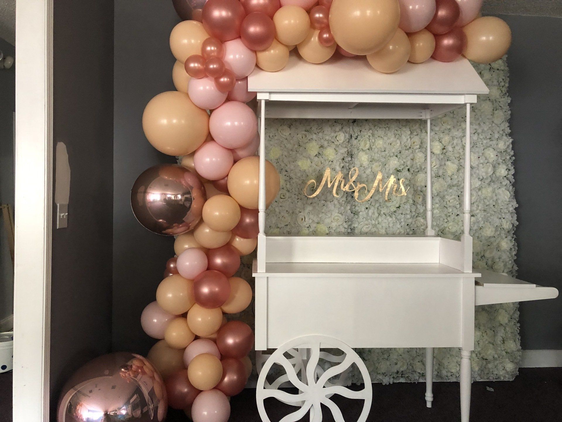 Blush peach and rose gold balloon installation around white cart with floral wall backdrop