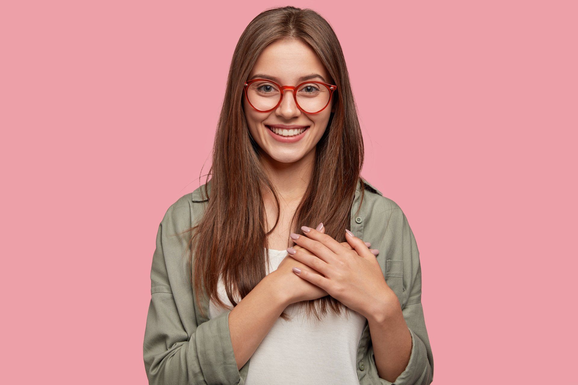 A woman wearing glasses is smiling and holding her hands over her heart.